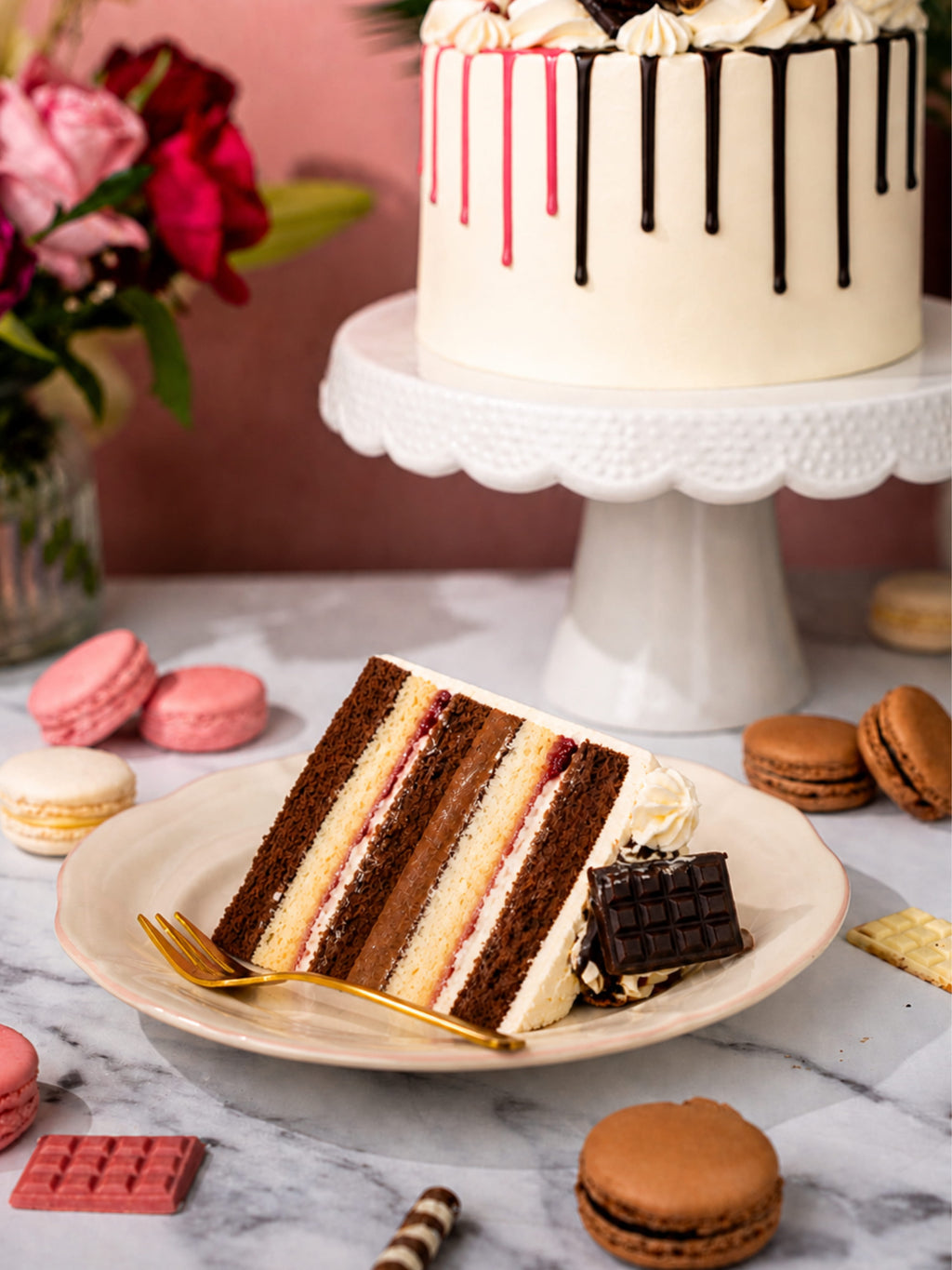 Slice of layered cake on a plate with a whole cake in the background, surrounded by macarons and flowers.
