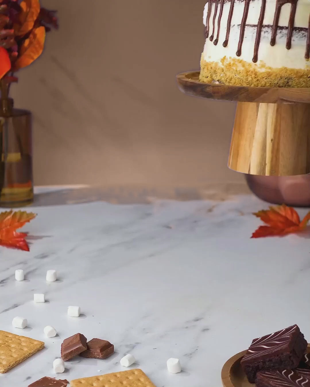 S'mores Cake with toasted meringue, brownie pieces and chocolate drip, followed by slicing and serving, against a warm autumn backdrop.