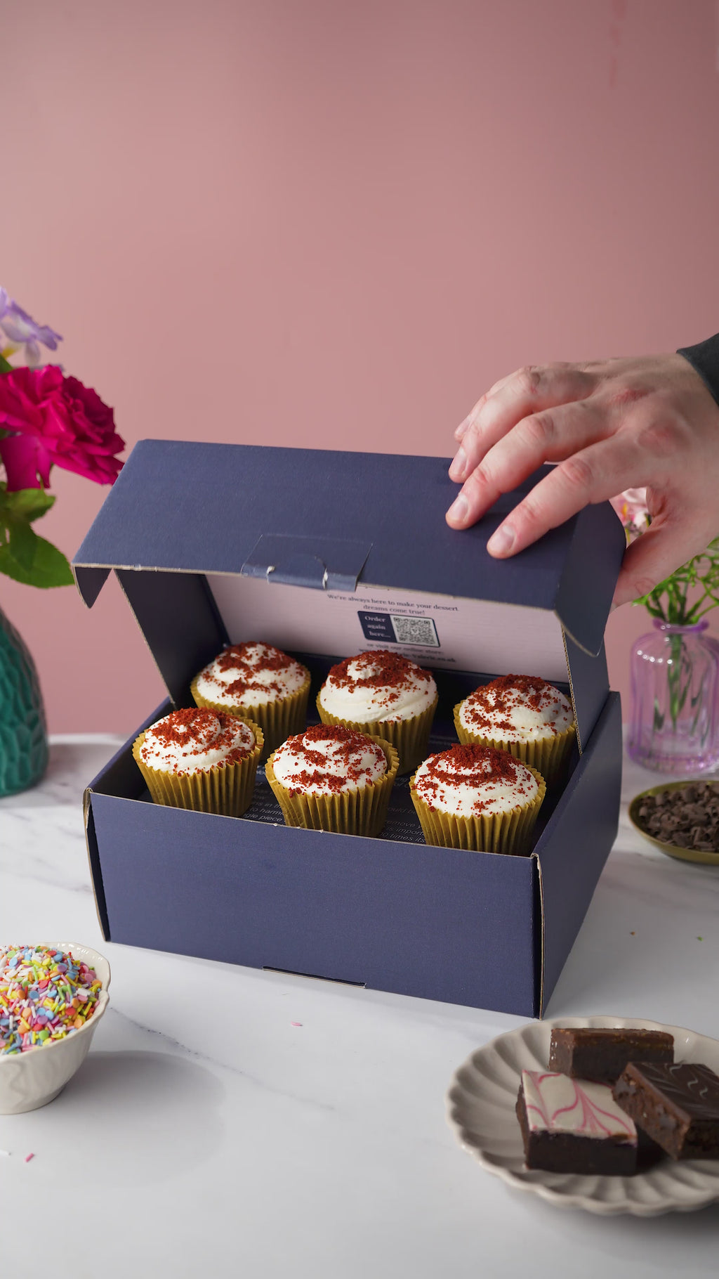 A box of red velvet cupcakes, with cream cheese frosting and a red velvet crumb topping. 