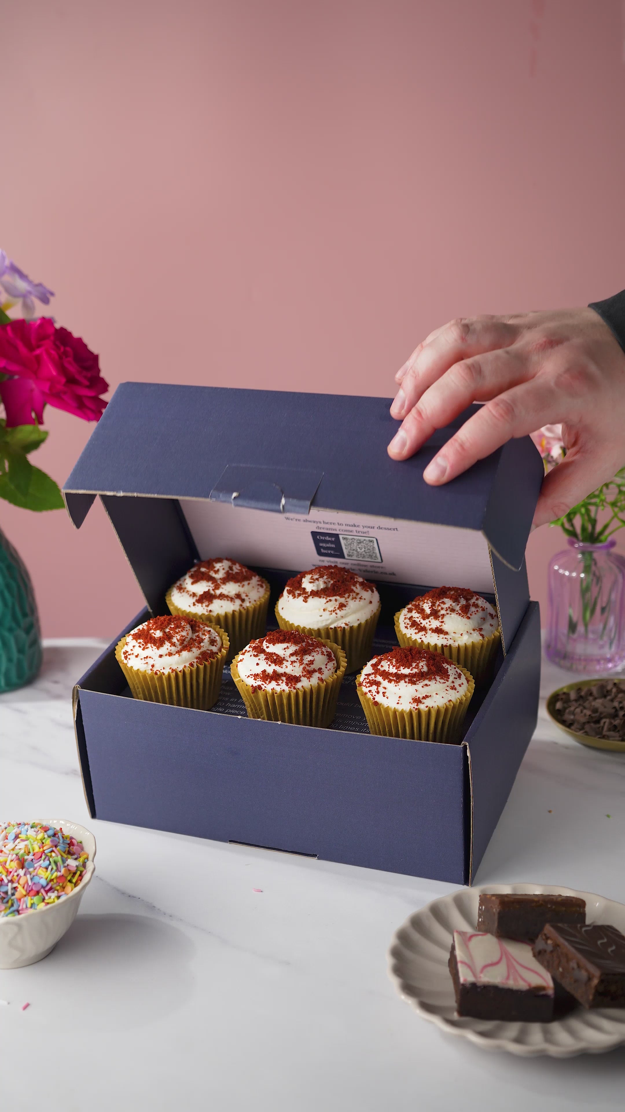 A box of red velvet cupcakes, with cream cheese frosting and a red velvet crumb topping. 