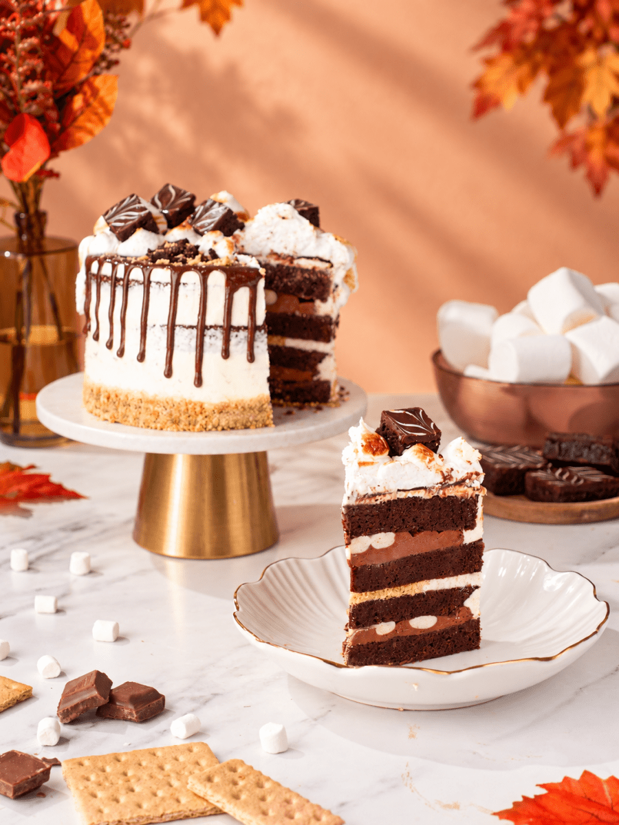 S'mores Cake with a slice removed and served alongside on a gold-rimmed plate, styled with autumn leaves.