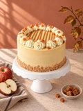 Toffee Apple Celebration Cake on a white cake stand with caramel drizzle, vanilla buttercream piping and fudge pieces